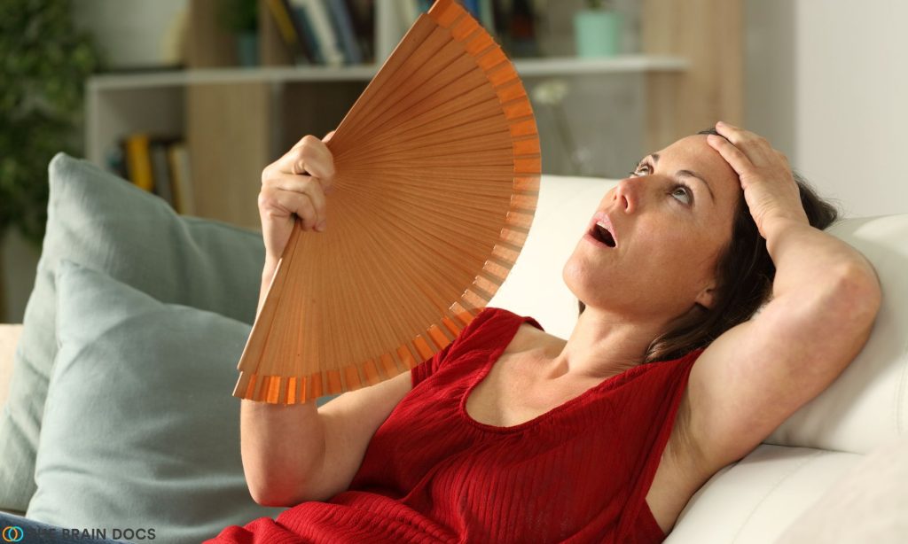 Menopausal woman with a red shirt sitting on a coach cooling herself with large hand fan.