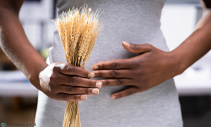 Woman holding bundle of wheat with gluten in foreground, in front of her stomach with her other hand on top.