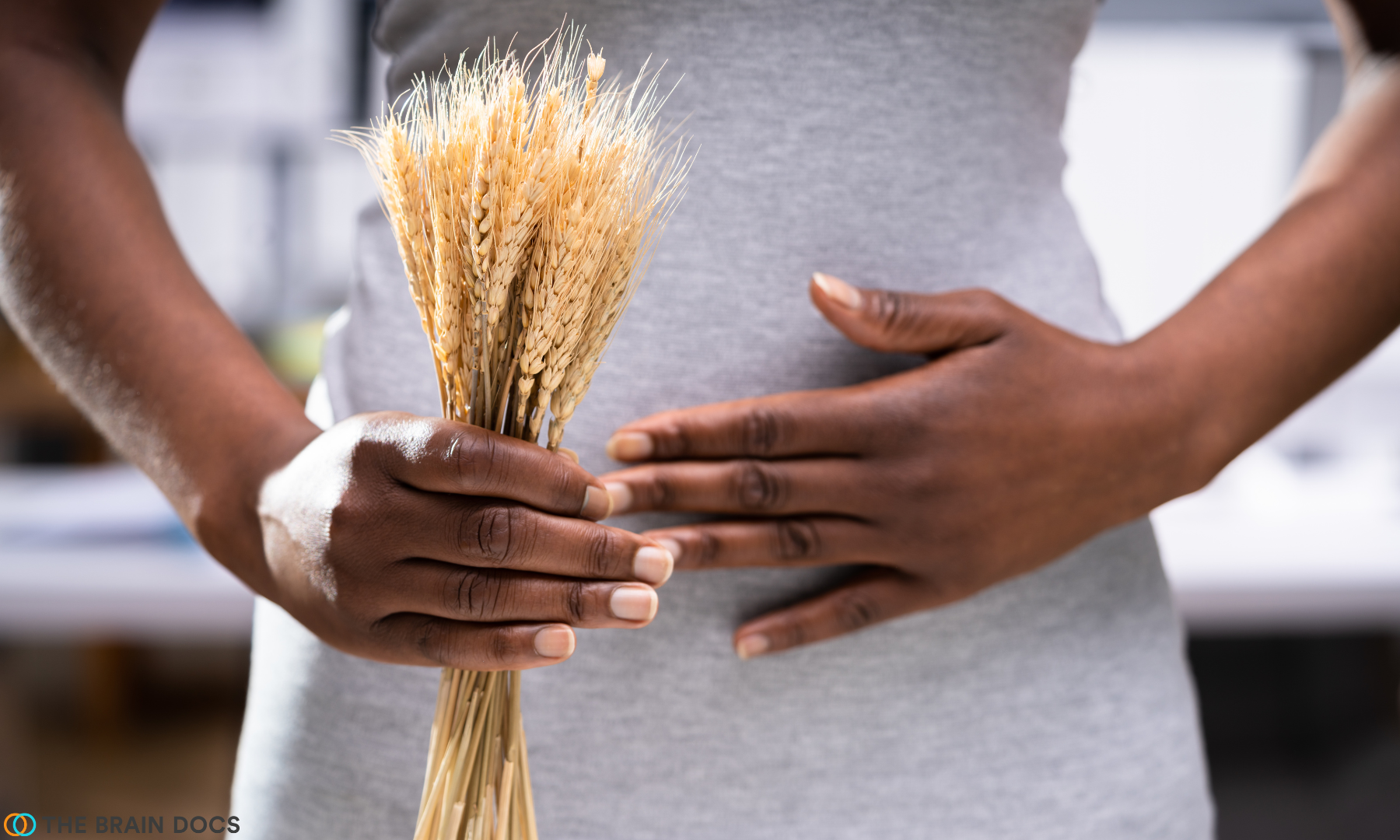 Woman holding bundle of wheat with gluten in foreground, in front of her stomach with her other hand on top.